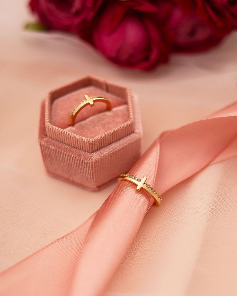 Anel dourado em estojo de veludo rosa, com outro anel sobre tecido acetinado e rosas ao fundo