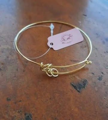Bracelete dourado fino com detalhe de nó e etiqueta rosa da E.M sobre mesa de madeira