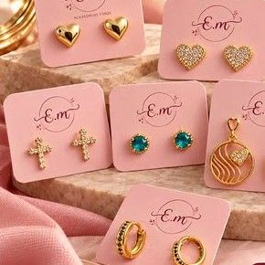 Brincos dourados da E.M em cartelas rosa: corações liso e cravejado, cruz cravejada, ponto de luz azul, mandala com coração e argolas com zircônias