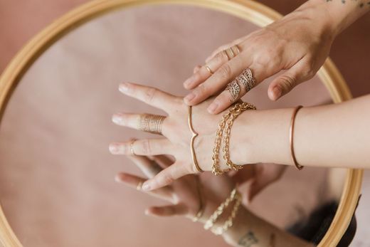 Mãos com anéis e pulseiras douradas refletidas em espelho redondo sobre fundo rosa